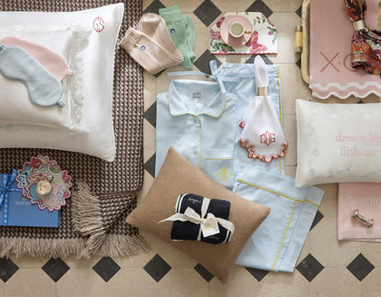 A styled flat-lay of baby bedding and nursery accessories on a patterned tile floor, including a light blue pajama set, white pillows with embroidered text, a pink sleep mask, beige socks, a small gift box tied with a ribbon, and other soft home textiles.
