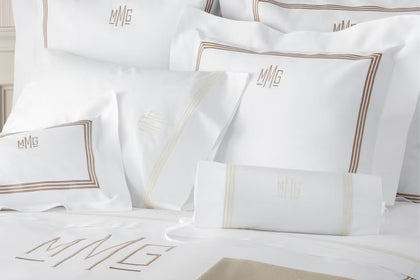 White bedding set with multiple pillows and a pillowcase, each decorated with gold embroidered borders and a matching 'MMG' monogram.