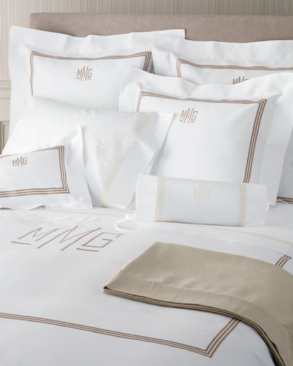 Bed dressed in white monogrammed bedding with tan embroidered lettering on the sheet, shams, and pillows, finished with a tan blanket.