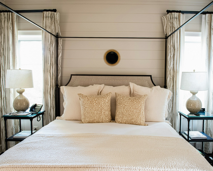 A beautifully styled bedroom featuring a four-poster canopy bed with cream bedding, two decorative beige patterned pillows, matching curtains, and two matching bedside tables with lamps.