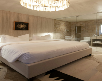 A modern bedroom featuring a large bed dressed in crisp white bedding, set against a light wood-paneled wall with a framed starburst artwork. A glass-enclosed bathroom is visible in the background, along with a hanging chandelier and a patterned rug.