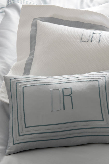 A close-up of white and light gray bedding with embroidered monogrammed pillows featuring the letters 'DR' in a muted blue-gray thread.