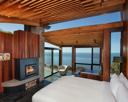 A luxury bedroom interior with a large bed in the foreground, a wood-burning fireplace, and expansive windows overlooking the ocean. The room features a slatted wooden ceiling, wood-paneled walls, and a modern open-concept layout.