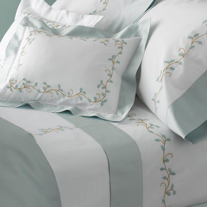 White bedding set with sage green satin borders and gold embroidered vine details, styled with matching pillows and a striped duvet cover.