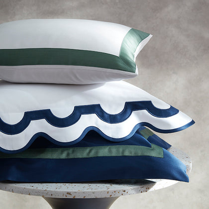 Three decorative pillows stacked on a round stone pedestal against a textured gray background. The pillows are white with contrasting satin trim in navy blue and sage green, featuring scalloped edges on the front pillow.