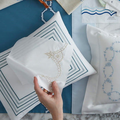 A hand holds a folded white embroidered fabric piece with a gold ornamental motif, surrounded by matching white pillows and blue patterned textile samples on a marble surface.