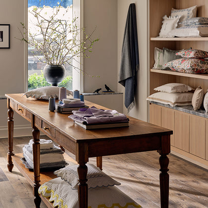 A warm-toned wooden dressing table in a styled bedroom or dressing room setting, surrounded by folded linens, pillows, shelves, and a window view.
