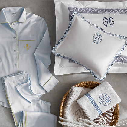 Light blue pajama set with matching pillowcases and a folded towel, arranged on a gray surface. The pajama top has gold piping and a gold embroidered crest on the chest pocket, and the bedding and towel are decorated with blue monogram embroidery and blue patterned borders.