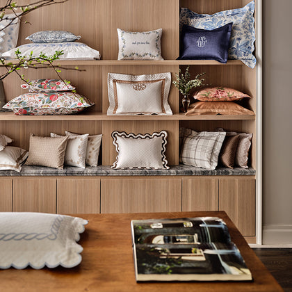 A styled interior scene showing a built-in wooden shelving unit filled with assorted decorative throw pillows in neutral tones, floral prints, and textured fabrics. A wooden table in the foreground holds a folded white pillow and an open magazine.