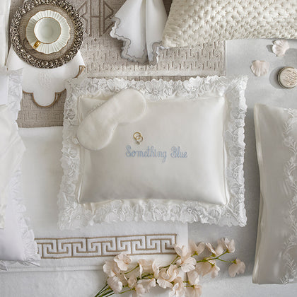 A styled bedding flat lay featuring a white satin pillow with a ruffled lace border, a matching sleep mask, and embroidered text reading 'Something Blue'. The scene is decorated with neutral-toned pillows, a teacup, and pale pink flowers.