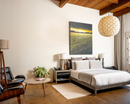 A modern bedroom interior with a bed dressed in white linens, a large vertical landscape painting above the headboard, a textured pendant light, wood ceiling beams, and a neutral rug on a wood floor.