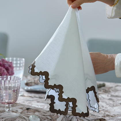 Hands unfolding Aziza white napkin with scalloped trim over Balthazar linen.
