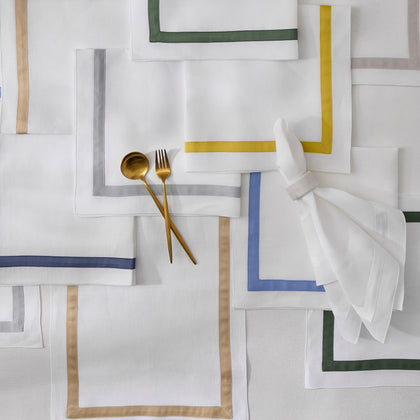 White Lowell napkins with bordered trim in assorted colors, arranged flat with gold flatware.
