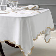 White linen tablecloth and placemats with a tan scalloped quatrefoil border, set with gold-rimmed glassware and a folded napkin, on a dark wood table.