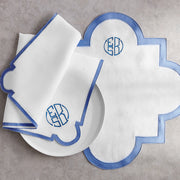 White linen scalloped quatrefoil placemat and napkins with a blue border and embroidered circular monogram, beside a white plate.