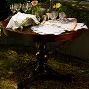 Round wood table set with scalloped quatrefoil placemats, stacked plates, napkins, wine glasses, and flowers, outdoors on a mossy patio.