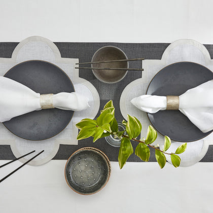 Overhead view of a table set with scalloped placemats, a dark runner, gray stoneware plates, napkins in metal rings, chopsticks, and small bowls.