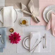 Overhead view of linen placemats and napkins with hemstitch borders in cream, tan, pink, and blue, china plates, teacup, cutlery, and flowers.