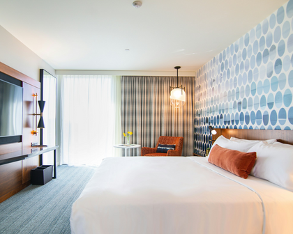 A modern hotel bedroom featuring a large bed with white linens and rust-orange accent pillows, set against a patterned blue and white accent wall. The room includes a wooden TV console, a small orange armchair, and a window with sheer white curtains and striped drapes.