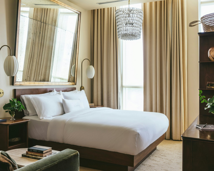 A luxury bedroom featuring a neatly made bed with crisp white linens, a dark wood frame, a large wall mirror, a hanging crystal chandelier, and floor-to-ceiling beige curtains.