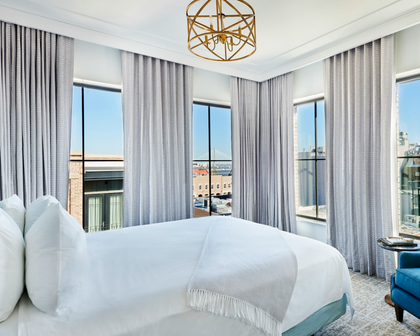 A modern bedroom interior featuring a neatly made bed with white linens and a fringed throw, large windows with light gray curtains, a gold geometric ceiling light, and a blue accent chair.