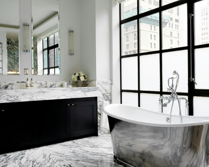 A luxury modern bathroom featuring a white freestanding bathtub with a chrome faucet, a black double-sink vanity with a white marble countertop and matching marble floor, and large black-framed windows looking out onto city buildings.