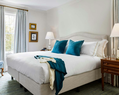 A styled bedroom featuring a bed with white bedding, two teal velvet pillows, a teal throw blanket, and a cream throw. The bed has a light beige upholstered frame, set against a wall with light blue curtains, a window, and a wooden nightstand.