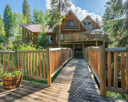 Triple Creek Ranch in Darby, MT: log cabin with wraparound deck reached by a rustic wooden footbridge through pines