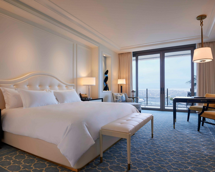 A luxury hotel bedroom featuring a large bed with crisp white linens, a tufted cream bench at the foot of the bed, blue patterned carpet, and floor-to-ceiling windows overlooking a scenic outdoor view.