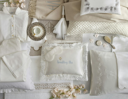 A styled flat-lay of a luxury bedding collection in soft ivory and cream tones, featuring a matching baby pajama set, textured pillows, lace-trimmed linens, a plush eye mask, and delicate floral accents.