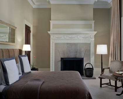 A styled bedroom interior featuring a bed with brown and gray bedding, a white fireplace mantel, two lit table lamps, a chair, and beige walls and curtains.
