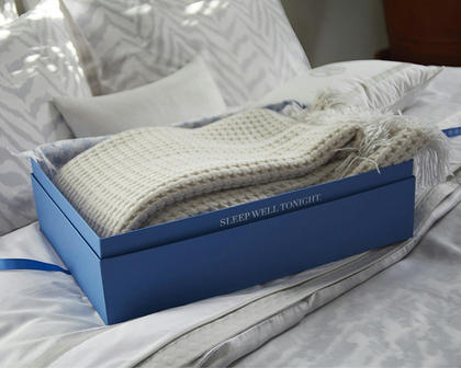 A folded cream knit blanket with fringe is presented in a blue box on a bed with white and gray patterned bedding. The box has the text 'SLEEP WELL TONIGHT' printed on the side.