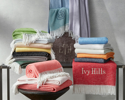 A styled product display of folded and rolled woven blankets and throws in multiple colors, arranged on black metal tables against a textured gray wall. One red throw with white fringe is embroidered with the name 'Ivy Hills'.