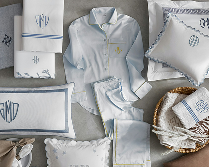 A styled flat-lay of a monogrammed men's bedding set in light blue and white, featuring a long-sleeve pajama shirt, matching pajama pants, pillows, and towels with blue and yellow embroidery, arranged on a gray textured background.