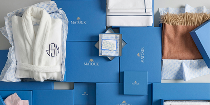 A luxury gift set from the brand MATOUK, featuring a white plush bathrobe with a monogrammed 'JSM', white towels, a patterned scarf, a brown fringed throw, and multiple royal blue branded gift boxes and cartons.