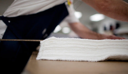 A close-up of a person in a white shirt and dark apron handling a folded white textured fabric or towel on a worktable.