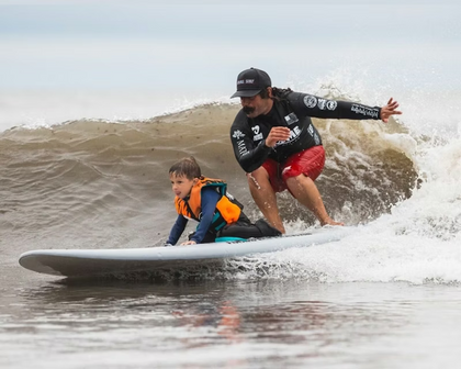 A surf instructor and young child riding a wave together, both wearing life vests.