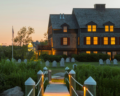 Weekapaug Inn in Westerly, RI at dusk, with shingled facade, Adirondack chairs on the lawn and a lit boardwalk path