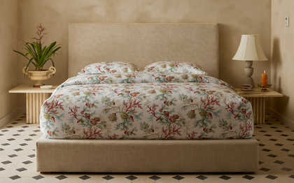 A bed dressed in a botanical print duvet cover and matching pillow shams, set in a styled bedroom with a beige upholstered headboard and footboard, flanked by wooden nightstands with a lamp and a potted plant.