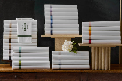 Stacks of folded white sheets with colorful ribbon trims beside a monogrammed guest towel and a white peony.
