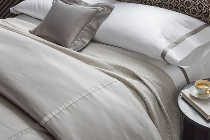 Bed styled with white hem-stitched sheets, a herringbone throw, and velvet pillows beside a nightstand with coffee and a book.