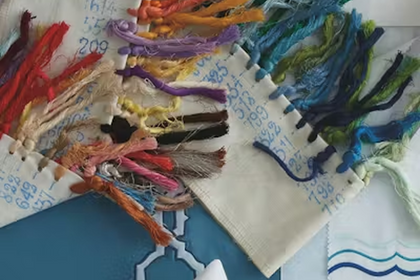 A close-up of a textile sample with multiple colorful tassels attached along the edge, photographed on a light surface with other fabric pieces partially visible.