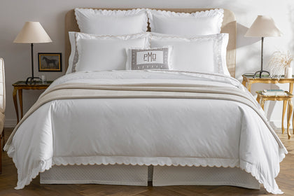 White scalloped bedding with a personalized monogram pillow, styled on a bed.
