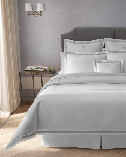 White monogrammed Ambrose sheets with a gray satin bordered trim, styled against a headboard.