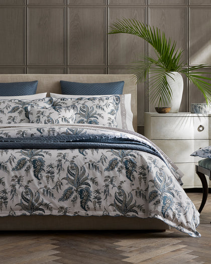 Blue-gray Apolline botanical print bedding with a quilted throw, styled on a bed.