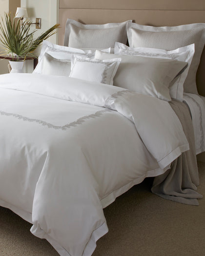 White Atoll bedding with pale blue coral-branch embroidered trim, layered on a bed beside a vase of flowers.