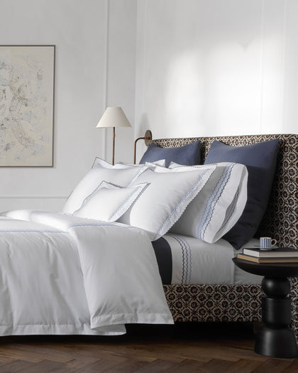 White Axel sheets with blue wavy embroidered trim and navy pillows, styled against a headboard.