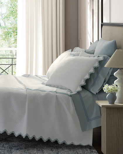 White Aziza Matelasse quilted bedding with a blue wavy trim, layered over blue sheets.