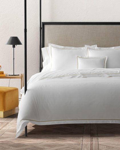 White Bergamo sheets with a gold single-stripe trim, styled on a bed beside an ottoman.