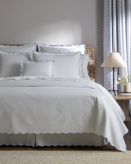 White Camilla Pique scalloped sheets with blue trim, styled against a headboard.